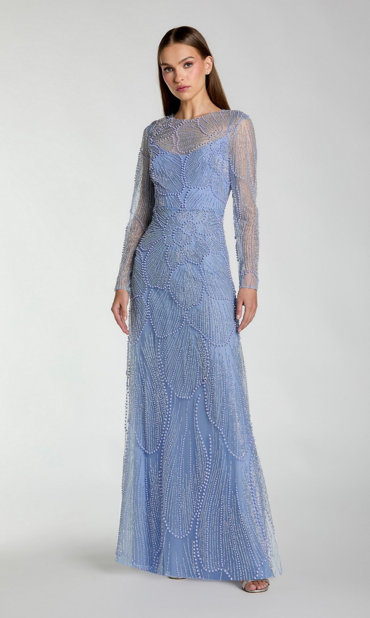 A woman wearing the Mac Duggal 69012 long formal dress—a fitted, periwinkle gown with sheer sleeves and intricate beaded embellishments—stands against a plain light background.